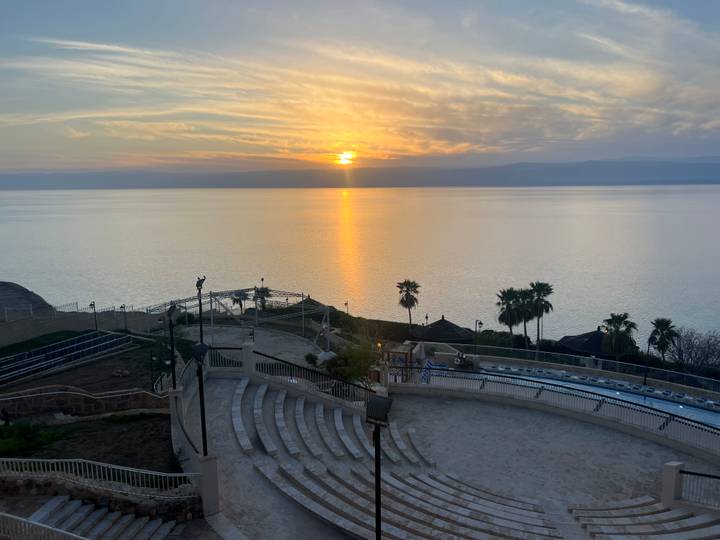 Golden sunset reflects over the tranquil Dead Sea with tiered seating and palm trees in the foreground.