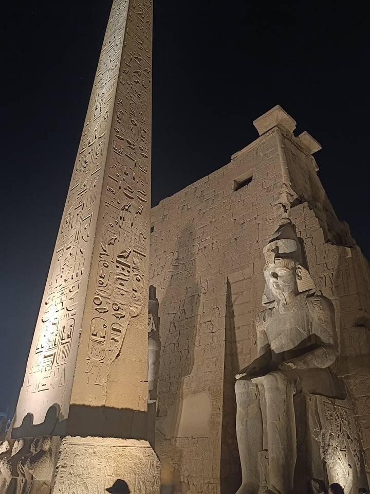 Illuminated Luxor obelisk and seated Ramses statue against dark night sky.