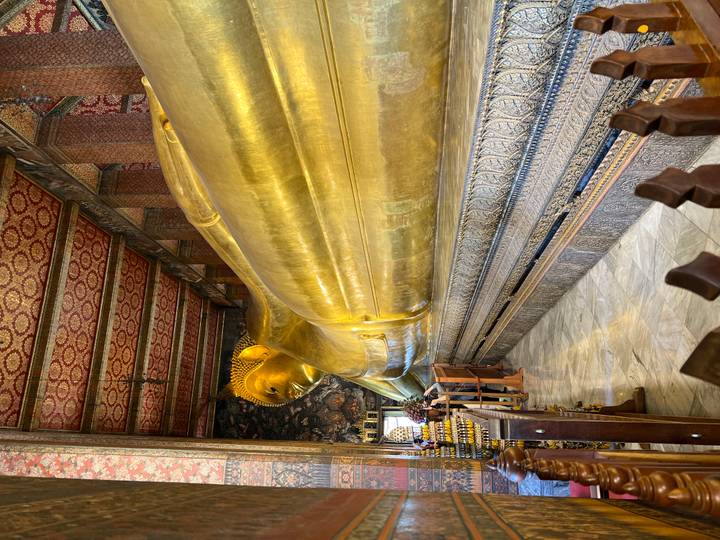 Gilded reclining Buddha stretches the length of an ornate temple hall decorated with patterned red ceiling.