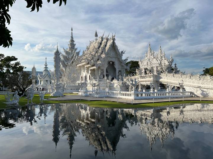 Dazzling white temple Wat Rong Khun gleams under bright sun, its reflection mirrored in a tranquil pond.