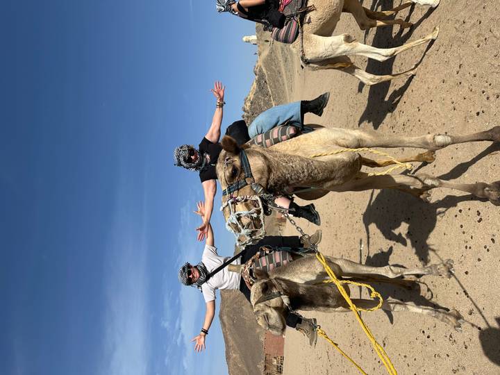 Two travellers riding camels with arms outstretched in a sandy desert landscape