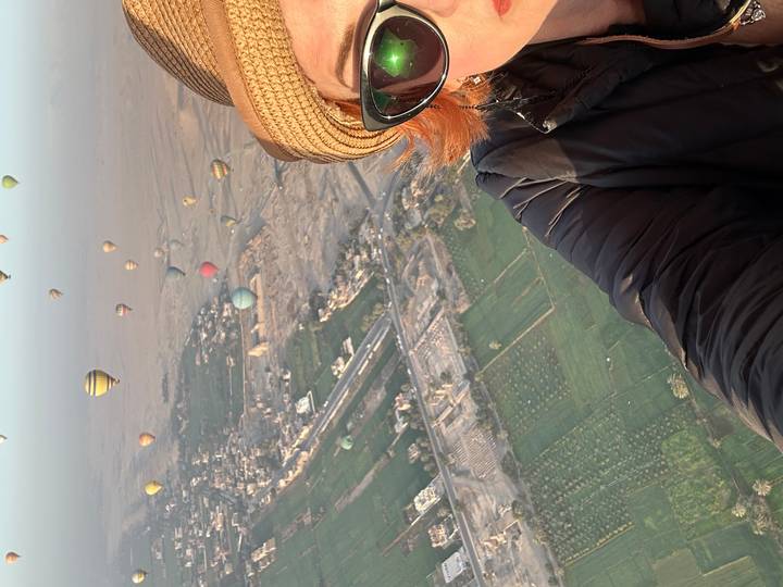 Self-portrait from a hot-air balloon overlooking green fields and dozens of colorful balloons at dawn