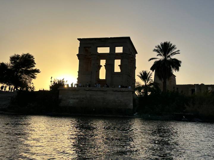 Silhouette of Philae Temple against an orange sunset with visitors standing along the columns by the Nile