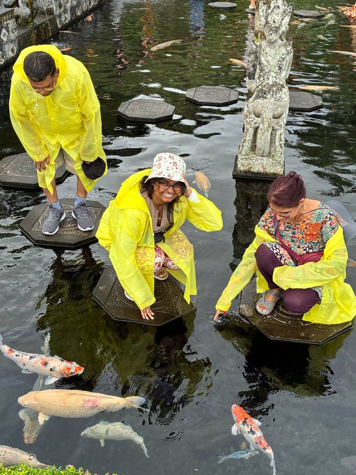 Two women in yellow rain ponchos balancing on stone stepping pads over a koi pond at Bali’s Tirta Gangga.