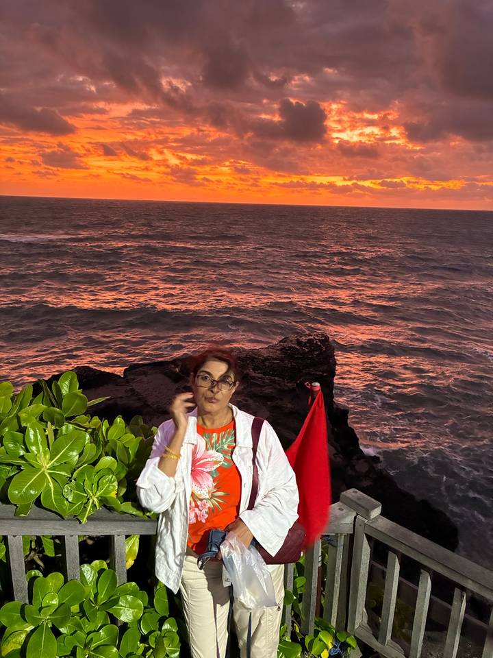 Lady posing by a rocky seaside cliff with dramatic orange sunset reflecting on the waves.