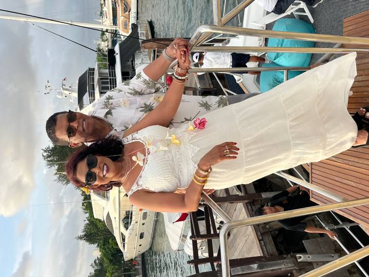 Smiling couple dressed in tropical attire on yacht boarding ramp with luxury boats behind.