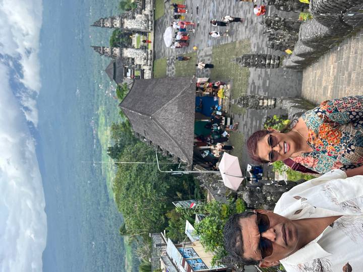 Smiling couple posing on stone steps of a Balinese hillside temple with lush valley and a cloud-capped volcano in the background