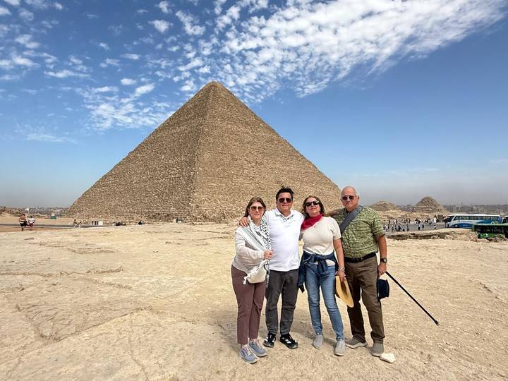 Four travelers stand smiling before the towering Great Pyramid under a bright blue sky with wispy clouds