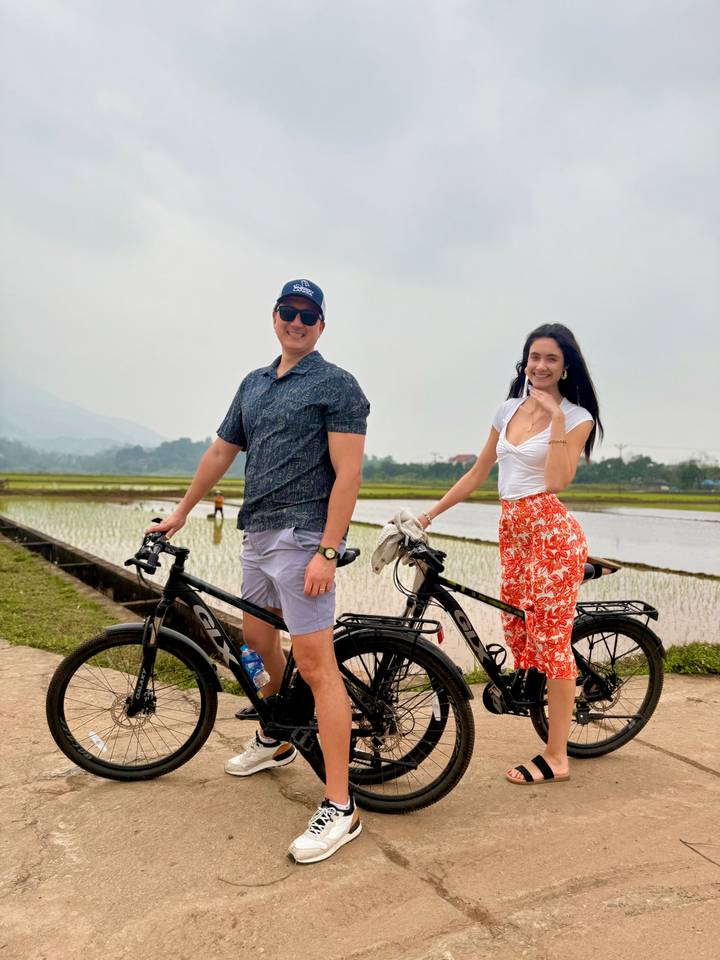 Couple on bicycles beside rice paddies under grey sky.