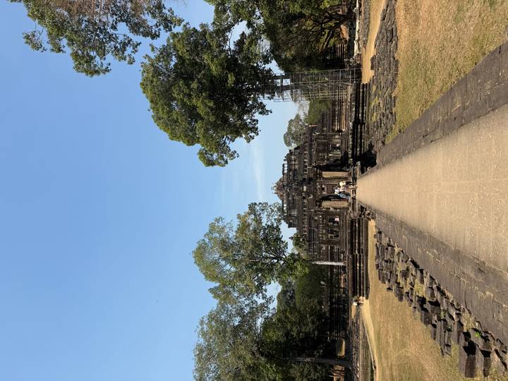 Long stone walkway leading to an Angkor era temple framed by trees and blue sky.