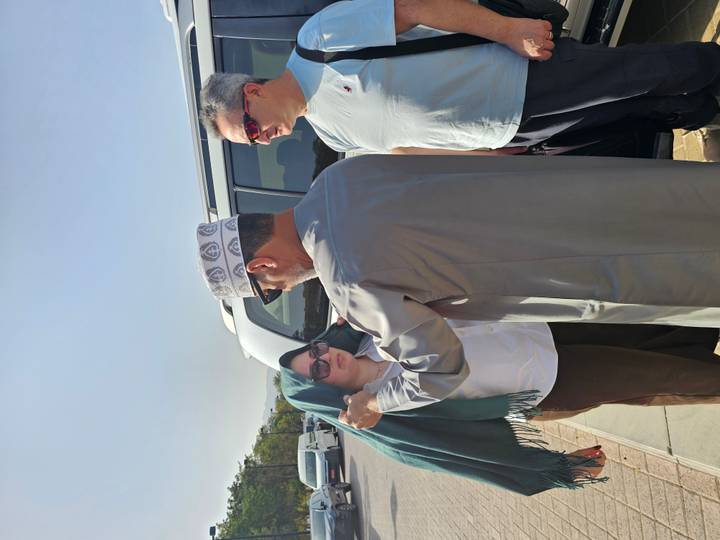 An Omani guide adjusts a headscarf on a female tourist beside an SUV while another traveler looks on under bright midday sun.