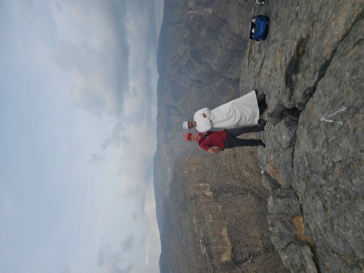 Two men stand on a rocky cliff edge overlooking a dramatic canyon landscape under a cloudy sky.
