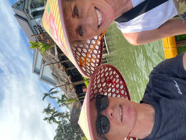Smiling couple wearing conical basket hats on a green canal with traditional houses behind.