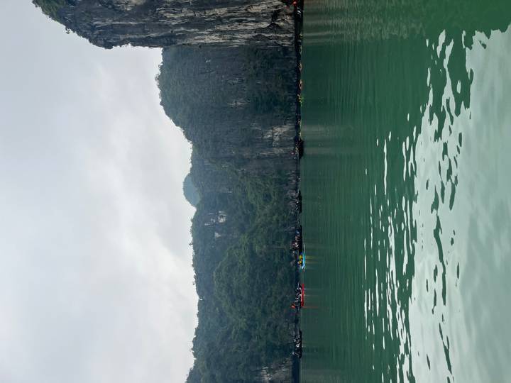 Calm emerald waters with distant kayakers and lush cliff faces beneath an overcast sky.