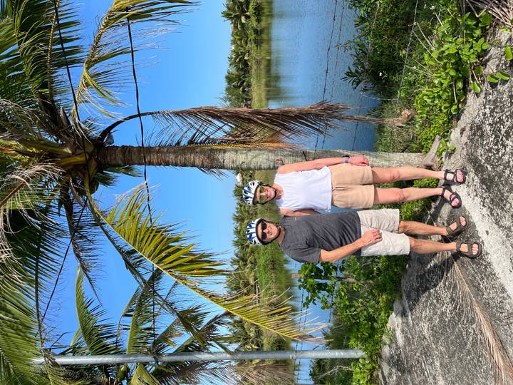 Couple in bike helmets stand by a lagoon and palm trees on a sunny day.