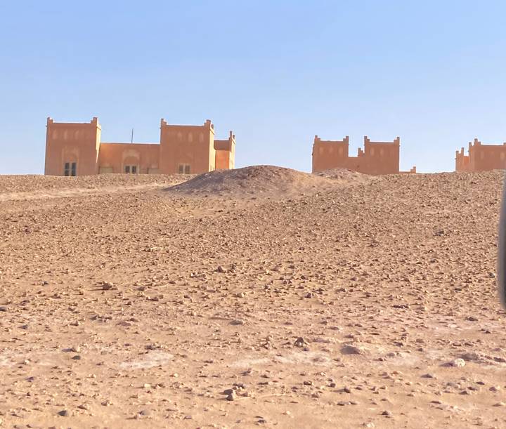 Isolated adobe-style buildings sit on a barren rocky desert plain beneath clear blue sky.