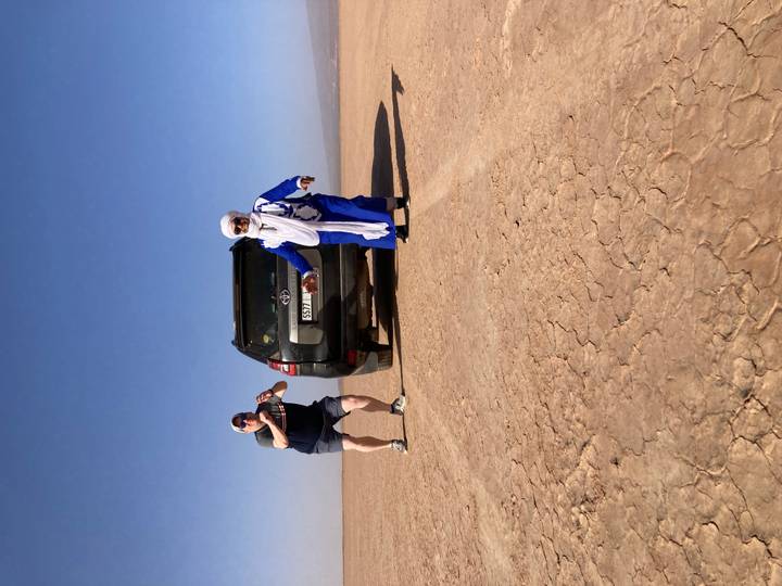Tourist and local guide pose beside 4x4 vehicle on cracked desert lakebed under bright sky.