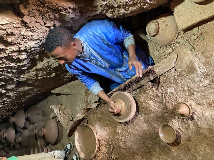 Man in blue robe shapes clay on a pottery wheel inside an earthen workshop.