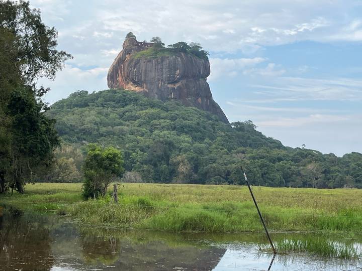 Iconic Sigiriya Rock Fortress rises above dense jungle and wetlands under a blue sky.