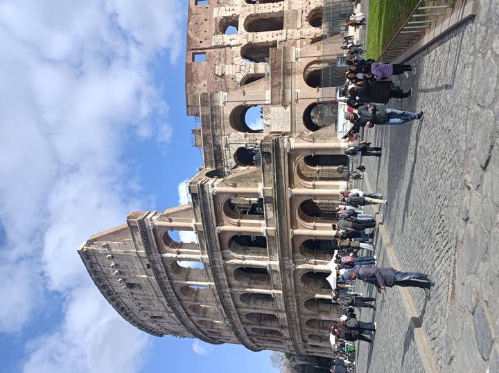 Street-level view of the Colosseum with many tourists walking on cobblestones under a blue sky with scattered clouds.
