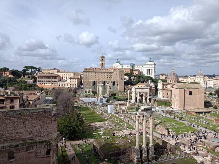 Panoramic overlook of the Roman Forum ruins framed by historic buildings and cloudy skies.