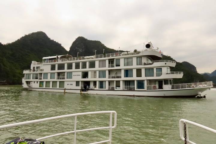 Modern white cruise ship gliding between forested limestone peaks on pale green water.