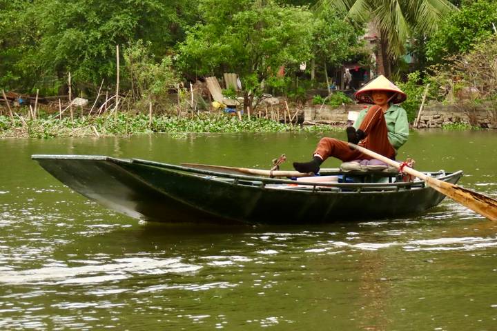 Person rowing a small metal skiff along a calm green river surrounded by lush vegetation.
