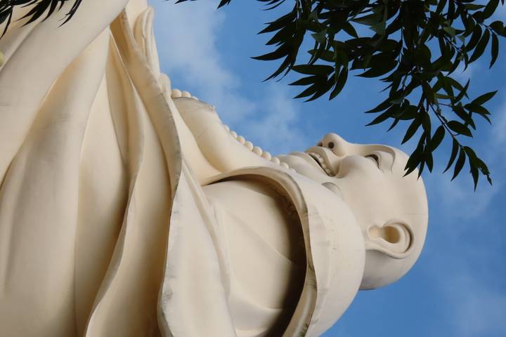 Large cream-colored smiling Buddha statue seen from the side against a blue sky.