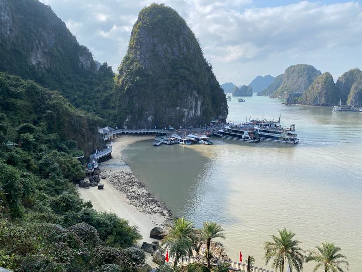 Panoramic view of a small sandy cove and pier surrounded by the limestone karst islands of Halong Bay with several cruise boats docked.