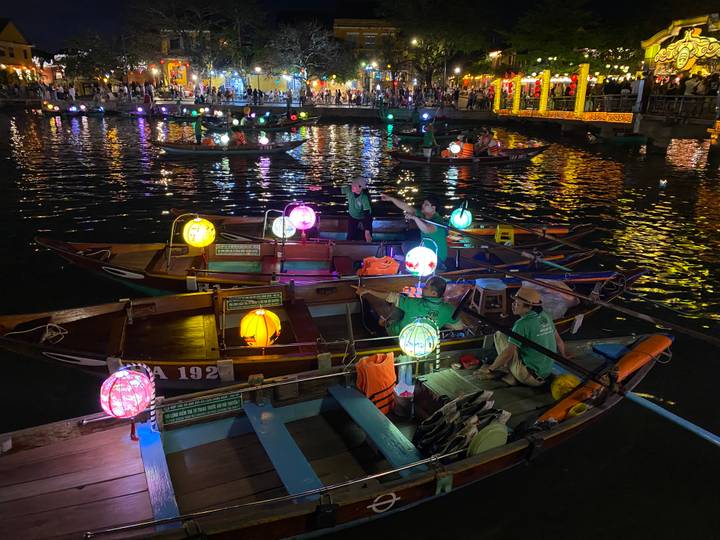 Colorful lantern-lit wooden boats on a river at night in front of a lively waterfront packed with people.