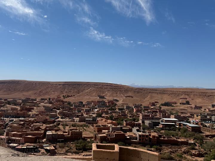 Brown adobe town sprawls across a desert ridge with high plateau and snowy peaks in background.