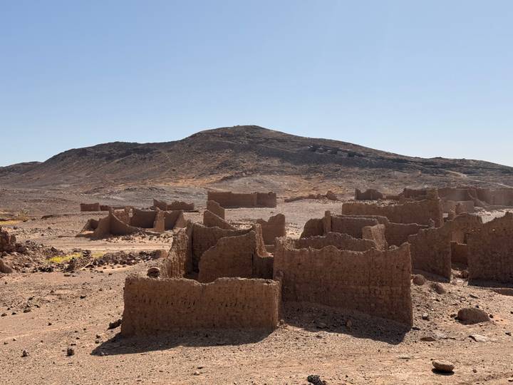 Crumbling adobe ruins scattered across a rocky desert plain beneath a barren hill.