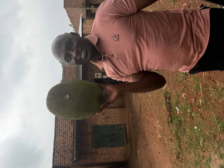 Smiling man in a polo shirt proudly holding up a large jackfruit outside a brick building.