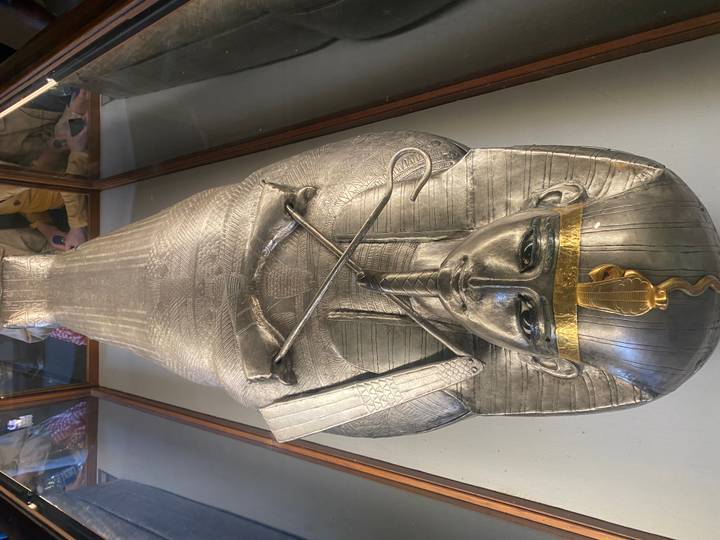 Silver sarcophagus displayed horizontally in a museum case with visitor reflections visible on the glass panels.
