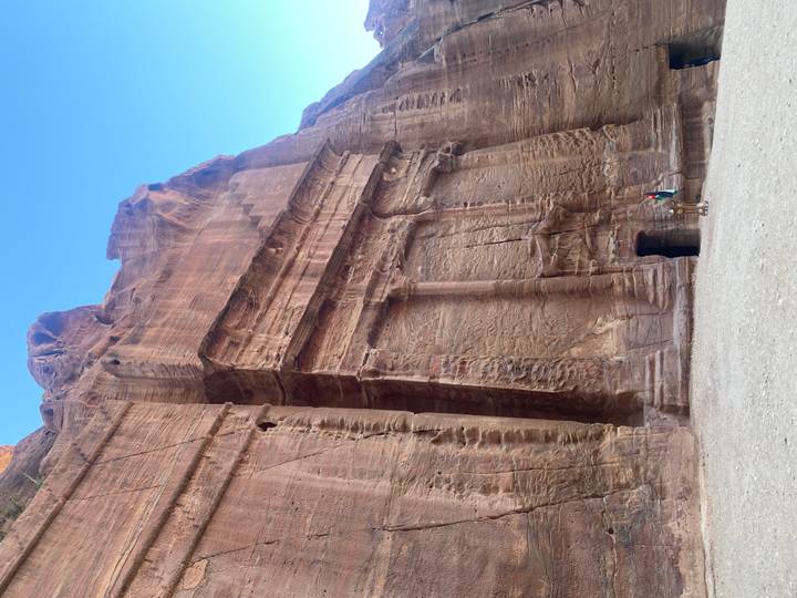 Massive sandstone façade in Petra with a single visitor standing at the entrance for scale beneath a clear sky.