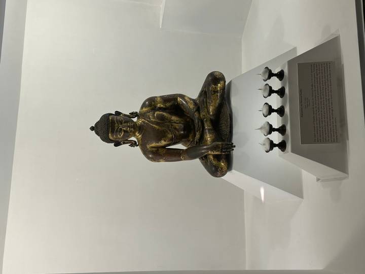 Bronze seated Buddha statue displayed in a minimalist museum case.