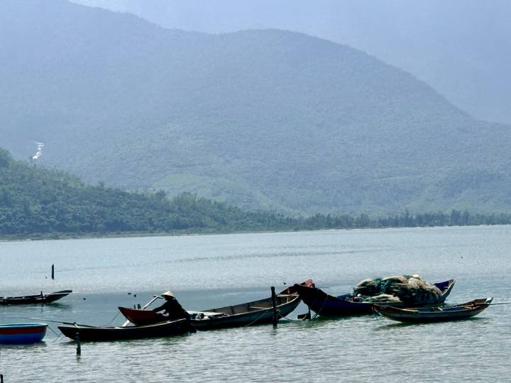 Small wooden fishing boats float on a calm lagoon backed by hazy mountains.