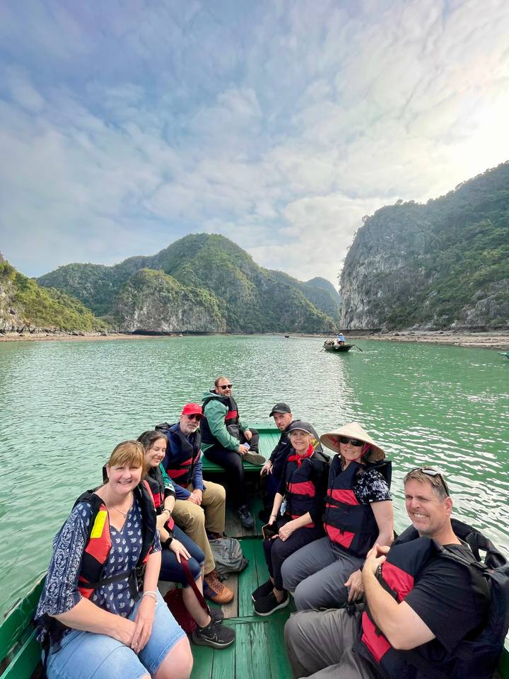 Travelers in life-jackets sit in a small boat surrounded by green karst cliffs and calm emerald water.
