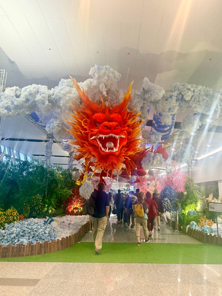 Indoor walkway filled with travelers beneath a dramatic red dragon decoration suspended among artificial clouds.