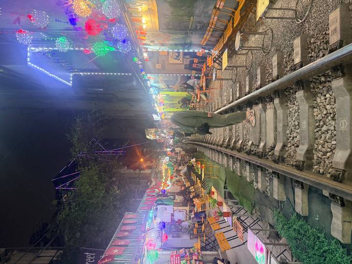 Traveler standing on narrow railway tracks flanked by lively night-time cafés and neon lights.