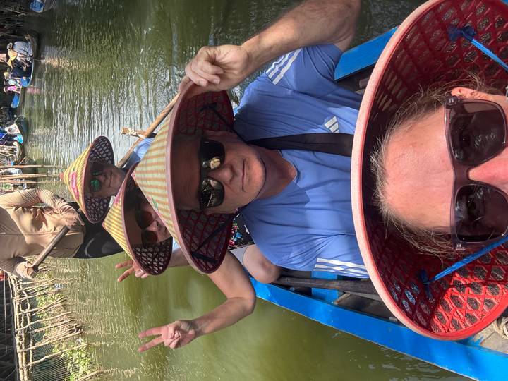 Group wearing conical hats smiles for a close selfie while rowing through a narrow, lush canal.