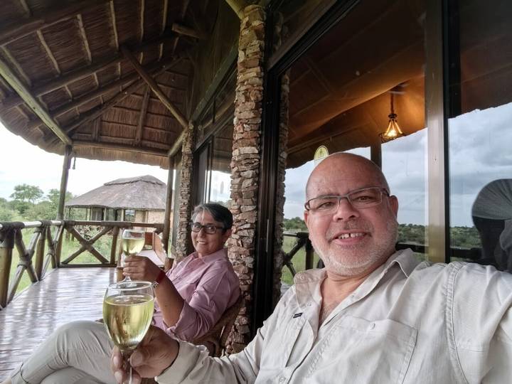 Two travelers enjoy glasses of wine on a stone-and-thatch lodge balcony overlooking the savanna.