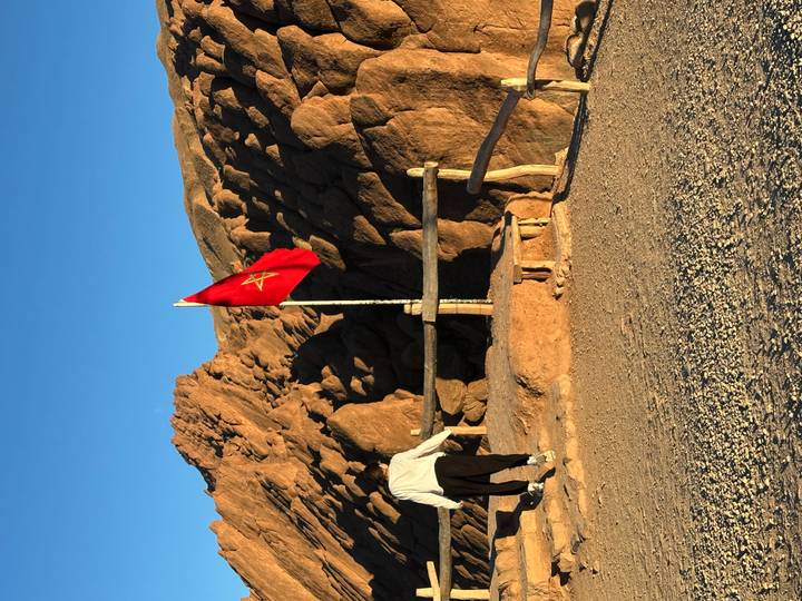 Hiker approaching a Moroccan flag on a rocky ridge under clear blue sky.