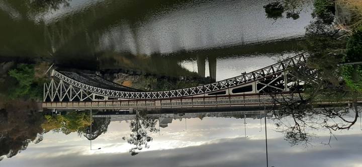 Ornate iron bridge spans a calm river beneath grey skies, reflections rippling in the water.