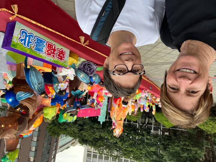 Two travelers smiling in front of a colorful outdoor lantern display.