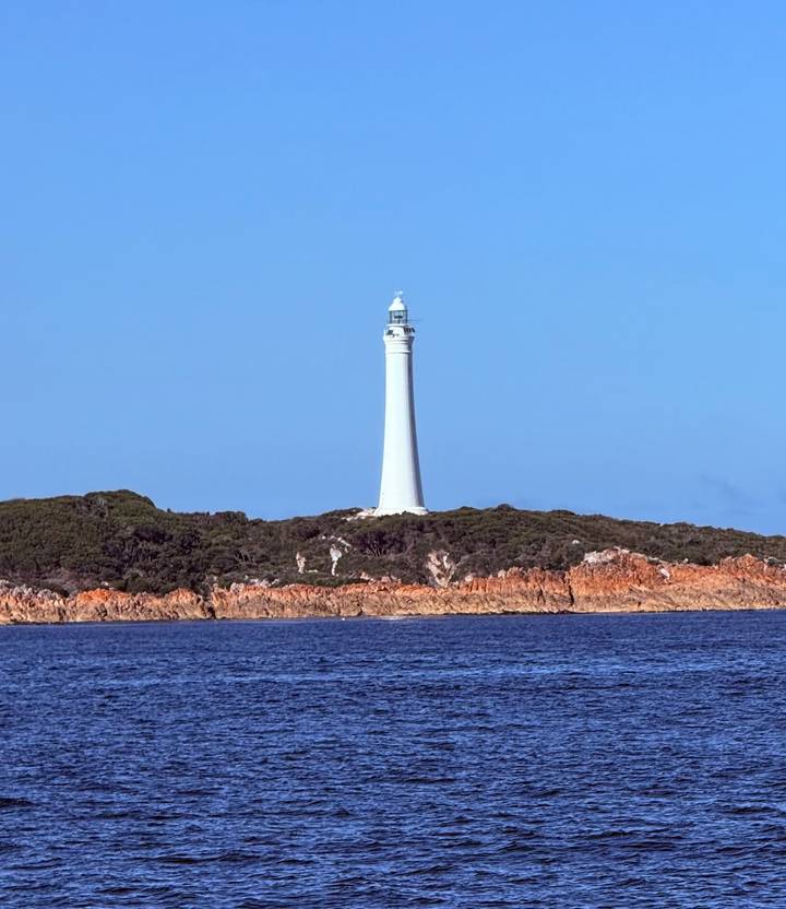 A solitary white lighthouse rises above a rocky, scrub-covered island against a clear sky.