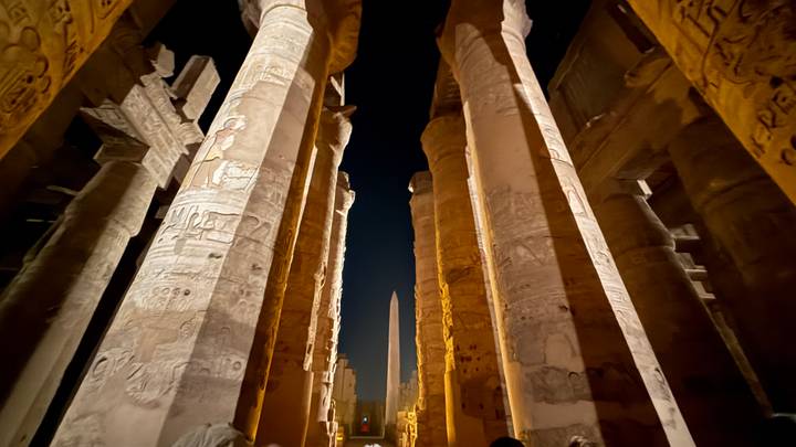 Night view looking upward between towering hieroglyph-covered columns toward an obelisk at Luxor Temple.