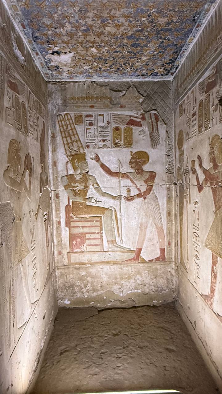 Painted tomb relief of a standing figure presenting a vessel to a seated pharaoh within an ancient chamber.