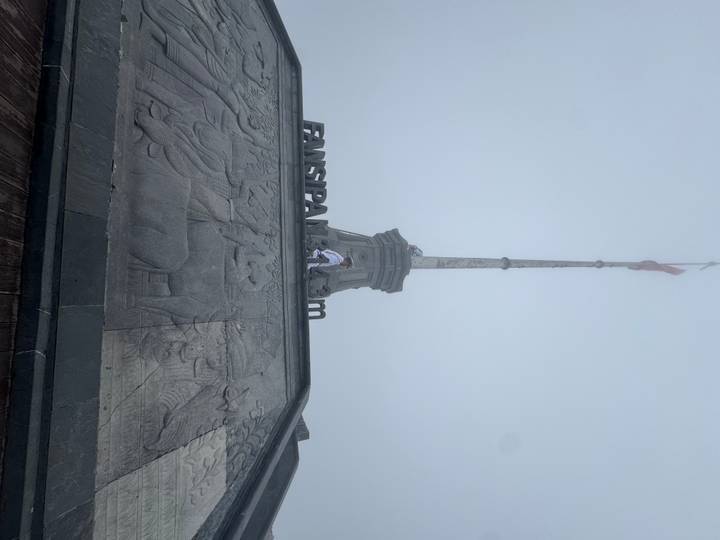 A foggy summit scene with a tall flagpole and engraved stone base at Fansipan 3,143 m.
