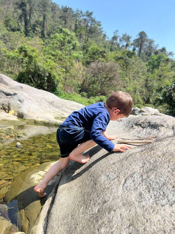 Young boy climbing over smooth river rocks beside clear shallow water in nature.